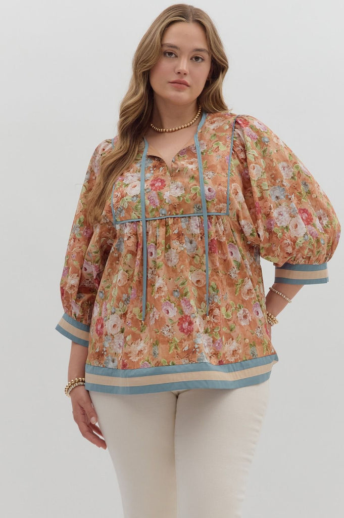 A woman wearing a loose-fitting floral blouse with puffed ¾ sleeves and an empire waist. The top has a vintage floral print in warm tones with blue and cream accent bands on the sleeves and hem.