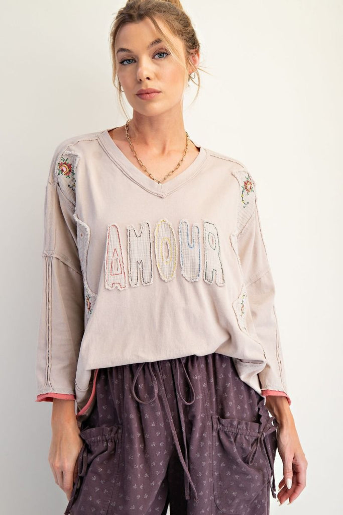 A woman wearing a mushroom-colored long-sleeve cotton jersey top with a V-neckline and drop shoulders. The shirt features appliqué letters spelling “AMOUR” across the chest, embroidered floral patch details on the shoulders and sides, exposed seams, and raw edges at the hem and sleeves. She pairs it with loose, purple drawstring cargo pants.