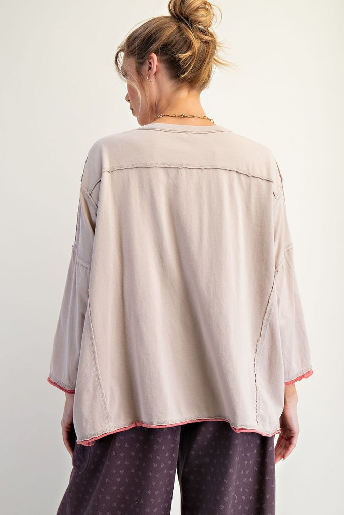 A woman wearing a mushroom-colored long-sleeve cotton jersey top with a V-neckline and drop shoulders. The shirt features appliqué letters spelling “AMOUR” across the chest, embroidered floral patch details on the shoulders and sides, exposed seams, and raw edges at the hem and sleeves. She pairs it with loose, purple drawstring cargo pants.
