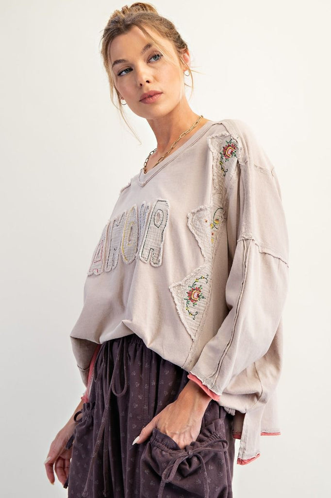 A woman wearing a mushroom-colored long-sleeve cotton jersey top with a V-neckline and drop shoulders. The shirt features appliqué letters spelling “AMOUR” across the chest, embroidered floral patch details on the shoulders and sides, exposed seams, and raw edges at the hem and sleeves. She pairs it with loose, purple drawstring cargo pants.