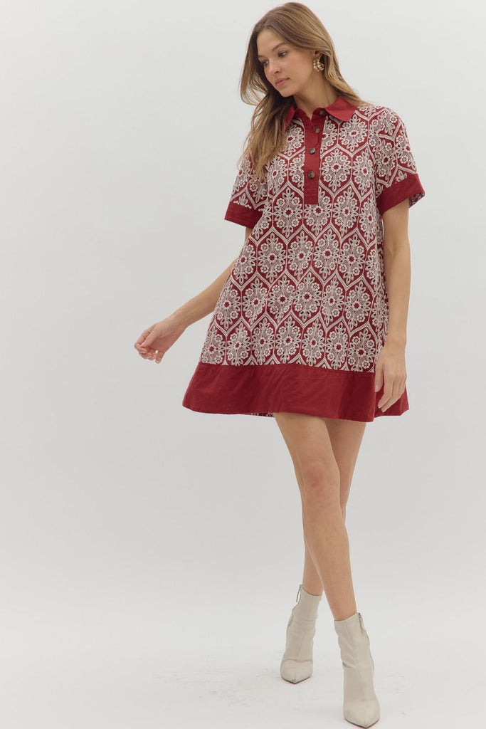 A short-sleeve swing dress in burgundy with a cream medallion-style embroidered print. It features a polo collar, a button-up placket, solid burgundy bands on the sleeves, and a wide solid burgundy hem, with side pockets.