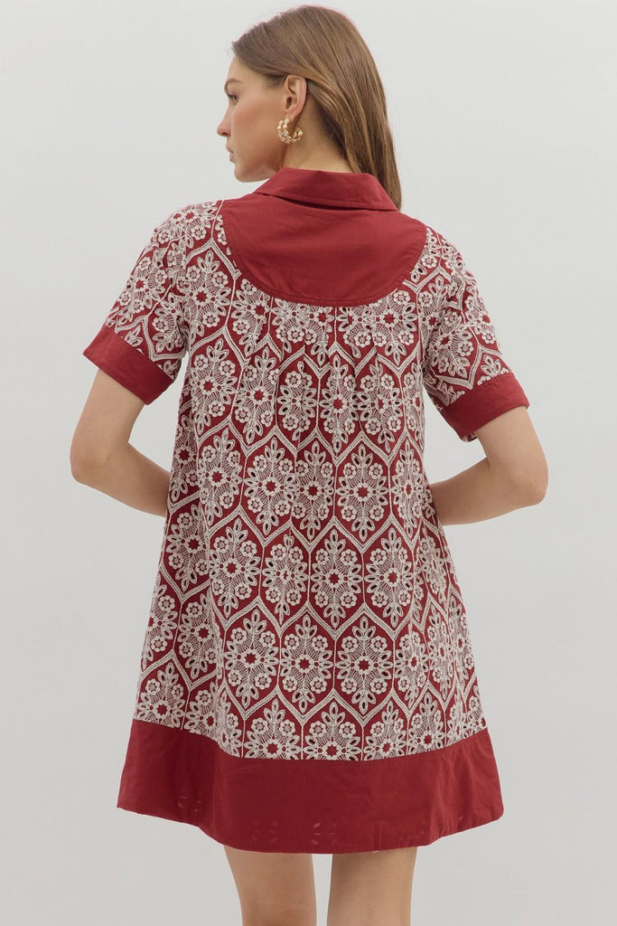 A short-sleeve swing dress in burgundy with a cream medallion-style embroidered print. It features a polo collar, a button-up placket, solid burgundy bands on the sleeves, and a wide solid burgundy hem, with side pockets.