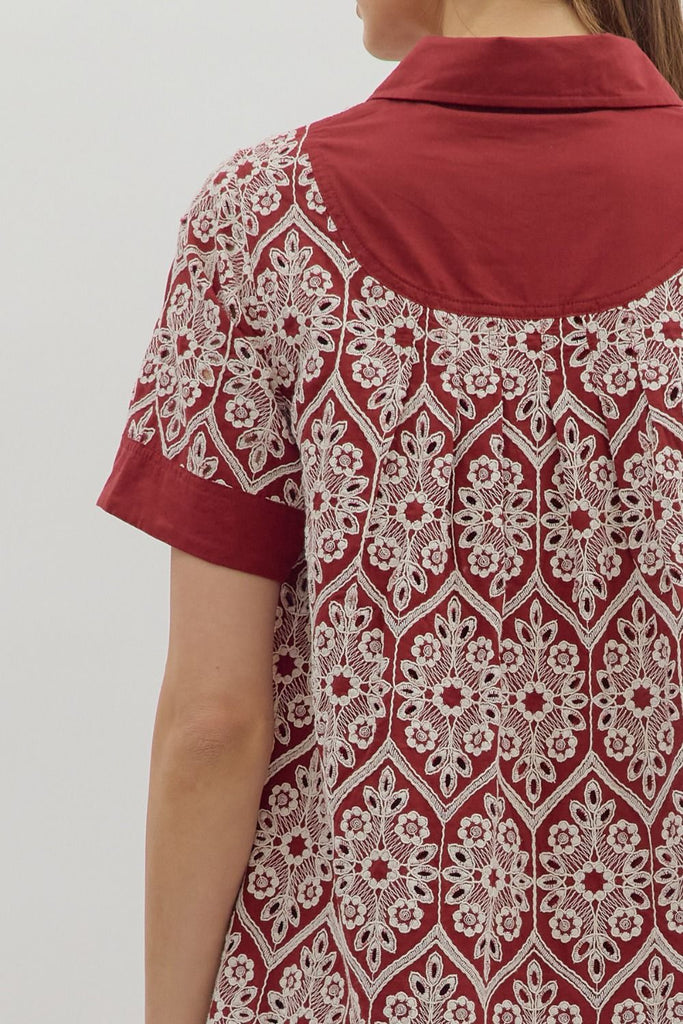 A short-sleeve swing dress in burgundy with a cream medallion-style embroidered print. It features a polo collar, a button-up placket, solid burgundy bands on the sleeves, and a wide solid burgundy hem, with side pockets.