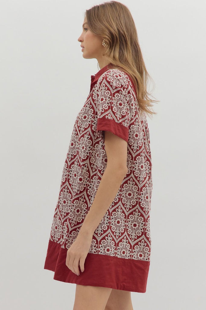 A short-sleeve swing dress in burgundy with a cream medallion-style embroidered print. It features a polo collar, a button-up placket, solid burgundy bands on the sleeves, and a wide solid burgundy hem, with side pockets.
