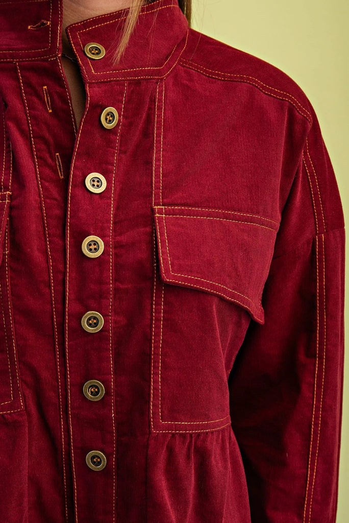 A burgundy corduroy jacket with gold contrast stitching, a button-up front with gold buttons, chest pockets, and a babydoll-style fit.