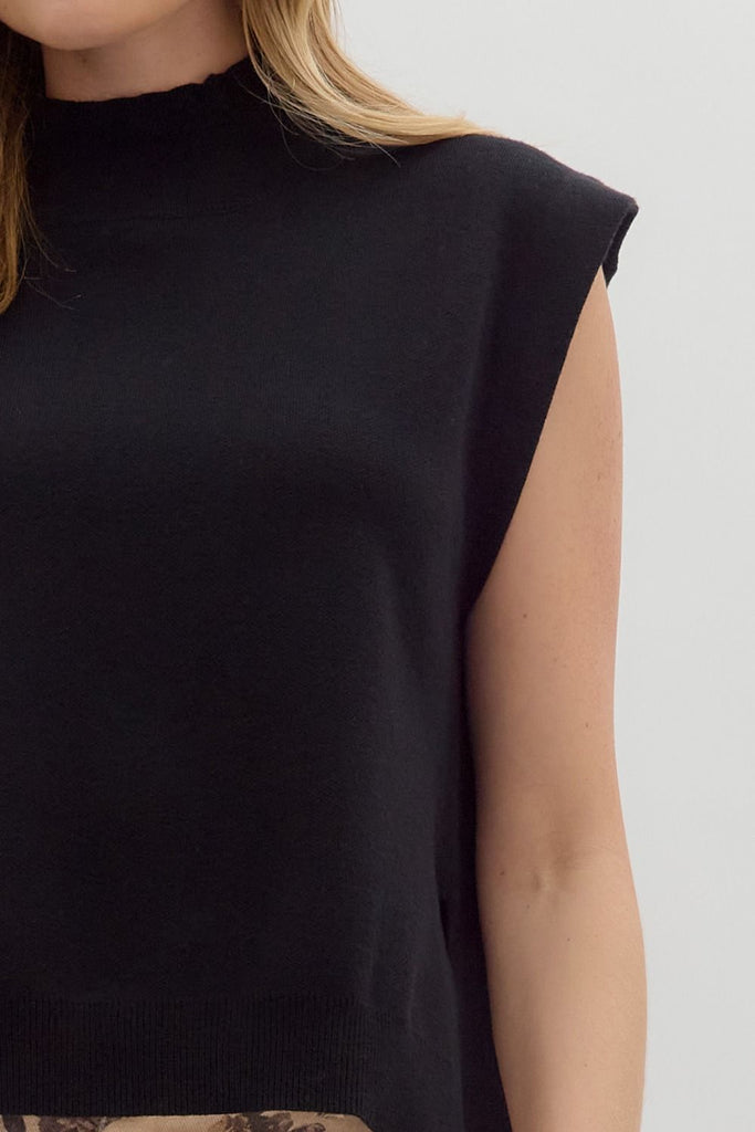 A woman models a solid black, sleeveless, mock-neck sweater with a high-low hem, paired with cute jeans.