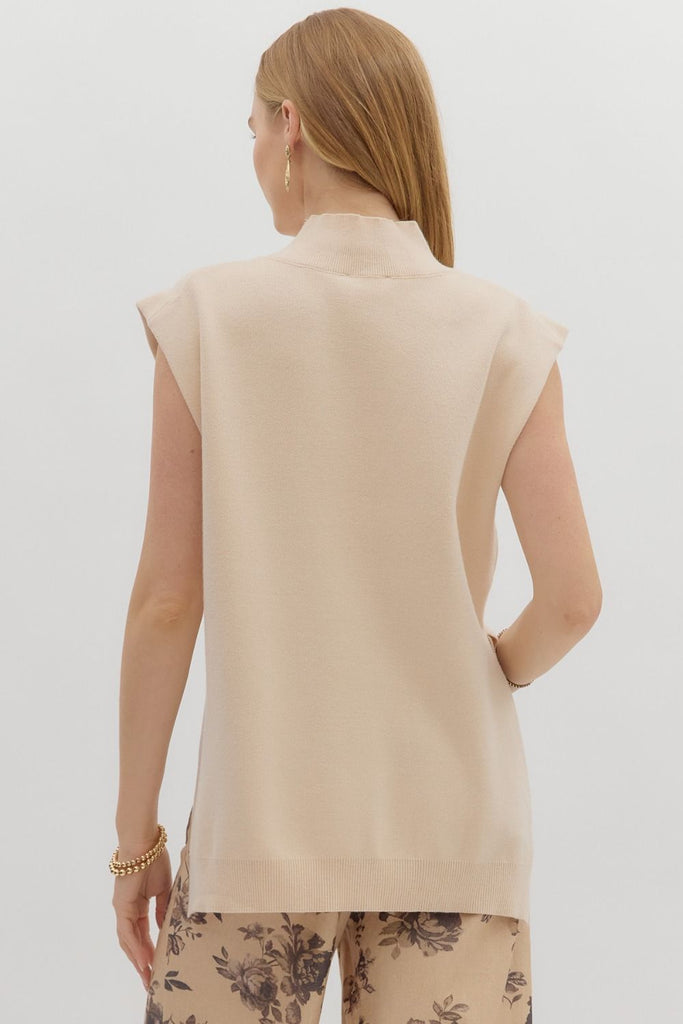 A woman models a solid oatmeal, sleeveless, mock-neck sweater with a high-low hem, paired with cute jeans.