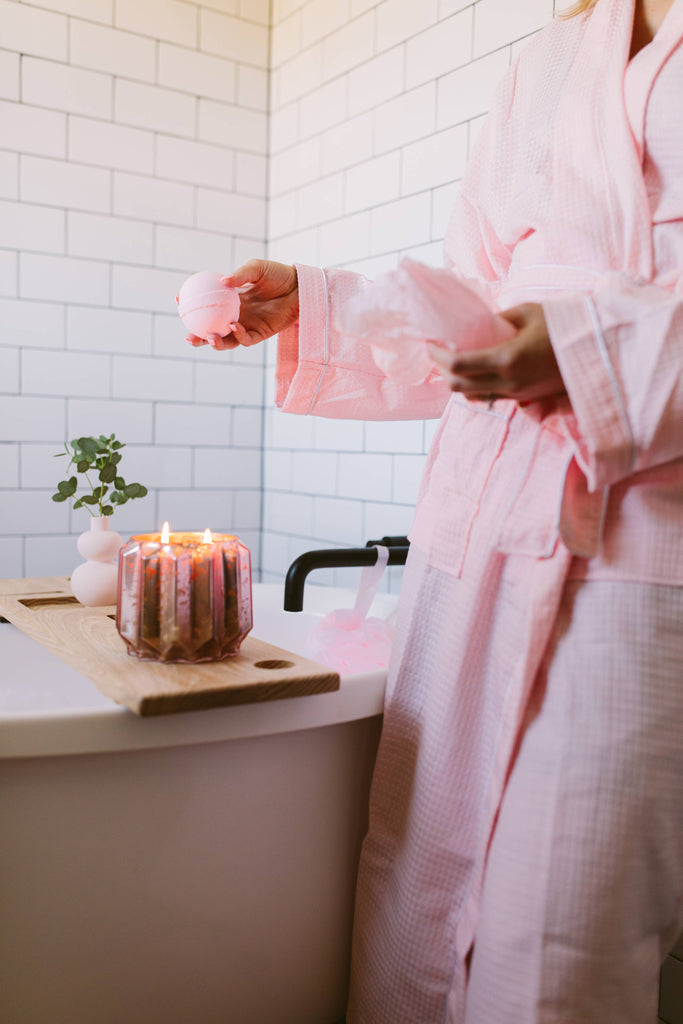 Person in a pink robe holding a candle in a bathroom setting.