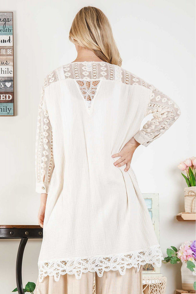 Woman wearing a natural-tone open cardigan with intricate white lace insets and scalloped hem, layered over a white ribbed crop top and beige wide-leg pants, standing indoors next to pastel floral décor.