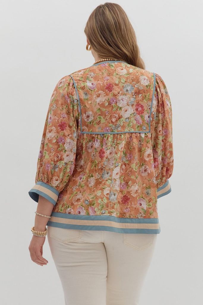 A woman wearing a loose-fitting floral blouse with puffed ¾ sleeves and an empire waist. The top has a vintage floral print in warm tones with blue and cream accent bands on the sleeves and hem.
