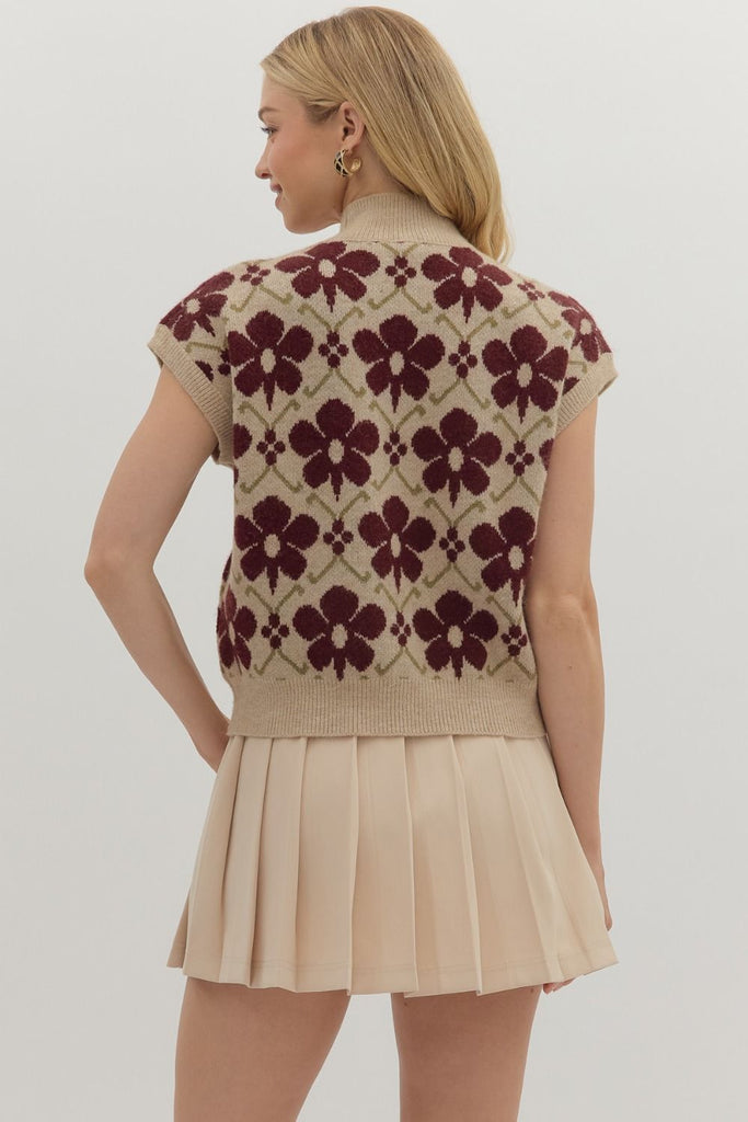 A woman wearing a sleeveless beige knit top with a burgundy floral pattern and high neckline, paired with a pleated beige mini skirt.