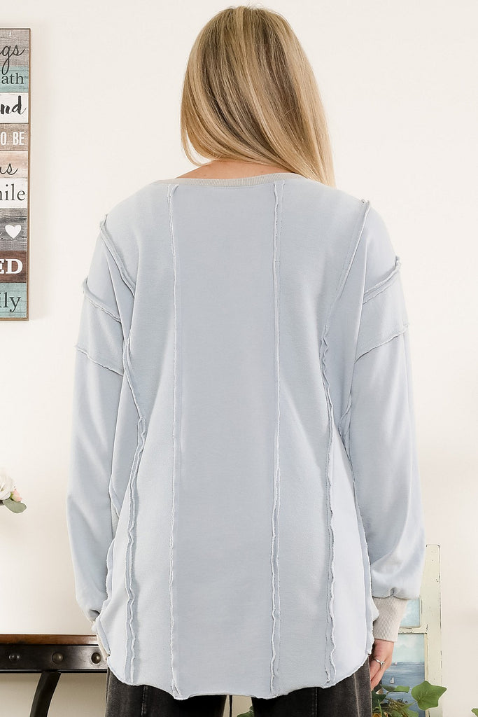 Smiling woman wearing a light blue washed terry sweatshirt with exposed raw seams and ribbed cuffs, styled with black jeans in a casual indoor setting.