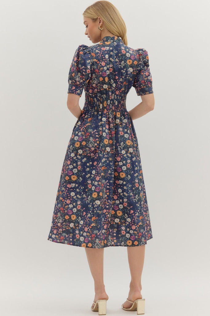 A woman wearing a navy midi dress with multicolor floral print, puff short sleeves, a smocked waist, and a front zipper. The dress features a flared skirt and side pockets.