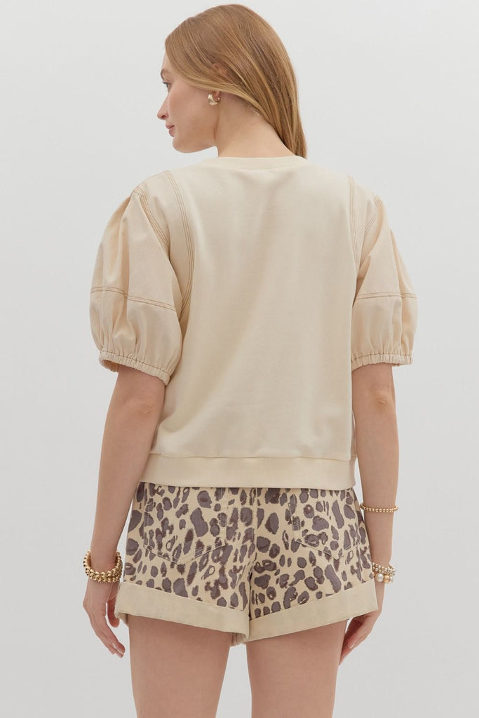 A woman wearing a beige crewneck top with short lantern sleeves and exposed seam details, paired with high-waisted leopard print shorts.