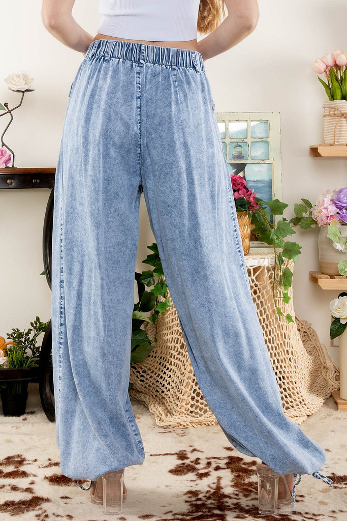 Close-up of a woman wearing high-rise, light-washed wide-leg denim pants with large snap-button patch pockets and adjustable drawstring cuffs, styled with clear high heels, standing indoors next to floral decor.
