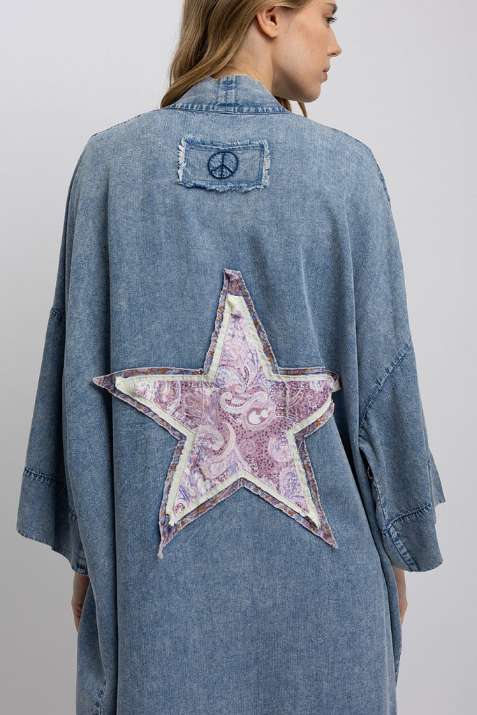 Back view of a model wearing a mid-length oversized denim kimono with a large paisley star patch and peace sign embroidery; front view shows fringe pocket details and relaxed open front styling.