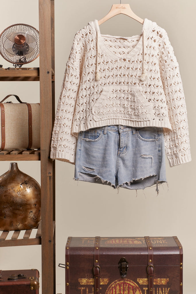 Beige crochet sweater and denim shorts hanging on a wooden rack with vintage suitcases and a fan in the background.