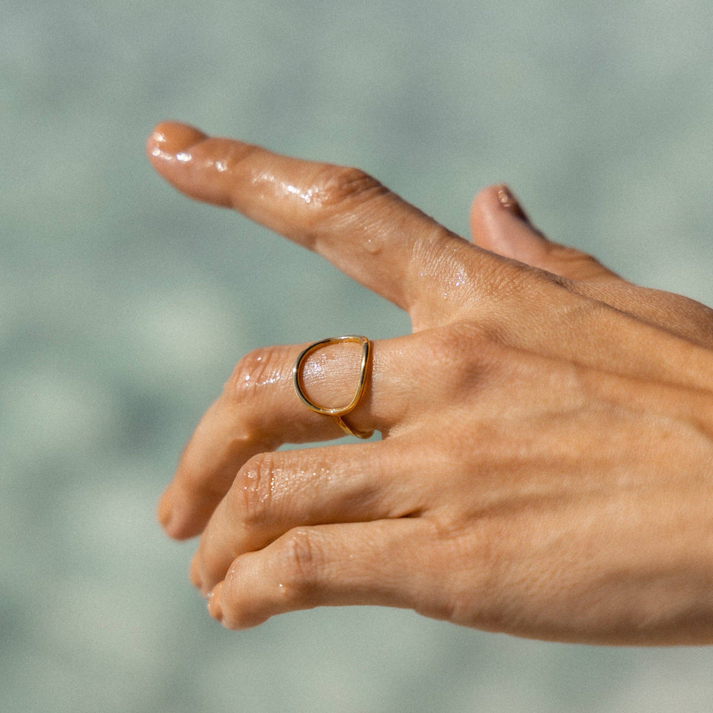 A close-up of a hand wearing a minimalist open-circle gold ring on the middle finger. The ring has a smooth, organic shape and a polished 18K gold finish, resting against sunlit, naturally textured skin.