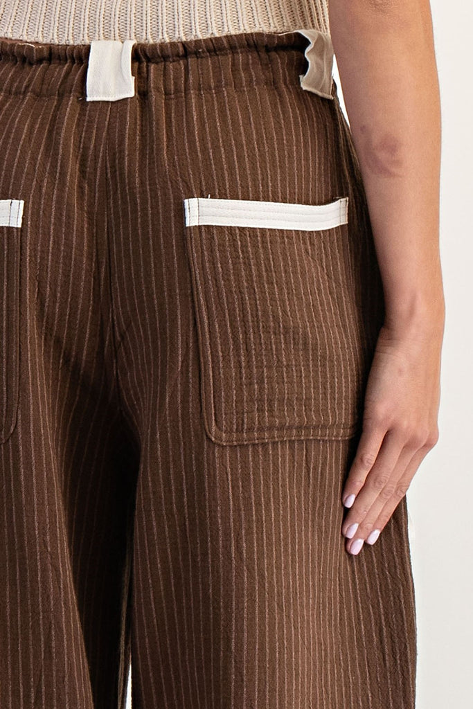 Model wearing relaxed-fit brown striped jogger pants with an elastic waistband and contrasting white drawstring ties. Pants feature utility-style flap pockets, front and back patch pockets, and gathered elastic hems, styled with open-toe logo slides.