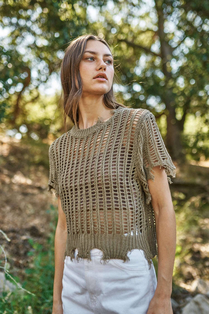 Woman wearing a brown knitted top and white pants standing in a natural setting with trees.