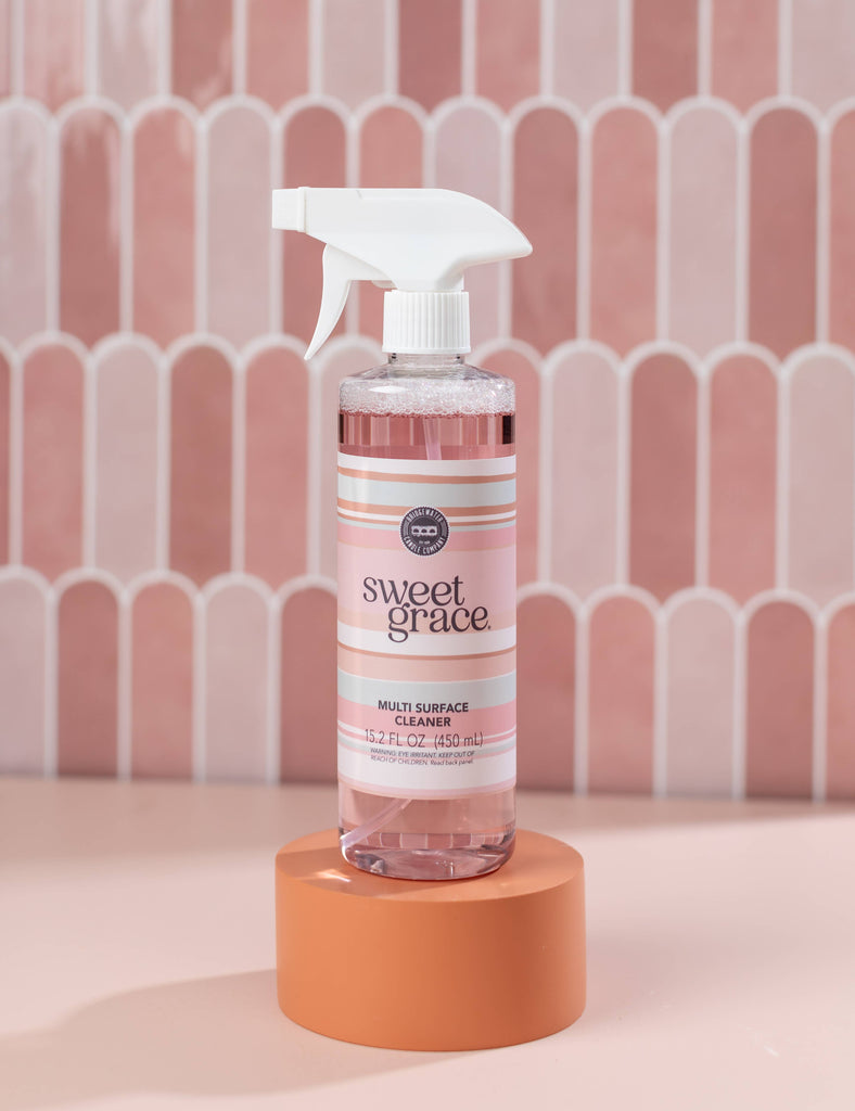 Bottle of Sweet Grace multi-surface cleaner on an orange stand with a pink geometric patterned wall in the background.