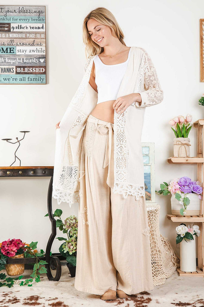 Woman wearing a natural-tone open cardigan with intricate white lace insets and scalloped hem, layered over a white ribbed crop top and beige wide-leg pants, standing indoors next to pastel floral décor.
