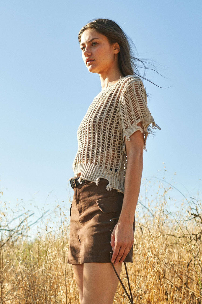 Woman wearing a beige crochet top and brown shorts standing in a field with a clear blue sky.