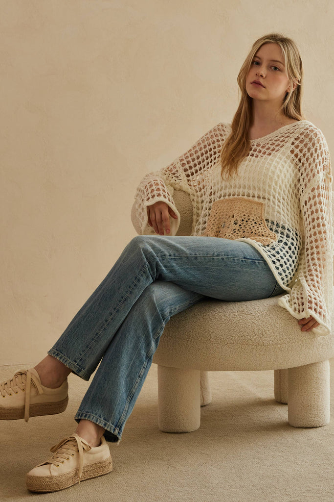 Woman sitting on a beige stool wearing a white knit sweater with a brown patch and blue jeans.