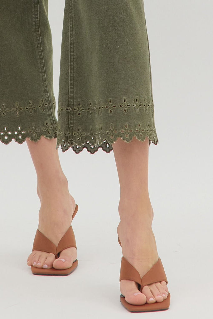A woman wearing olive green high-waisted, ankle-length pants with a scalloped hem featuring floral embroidery. The pants have a wide-leg fit and classic jean pockets, paired with brown heeled sandals.