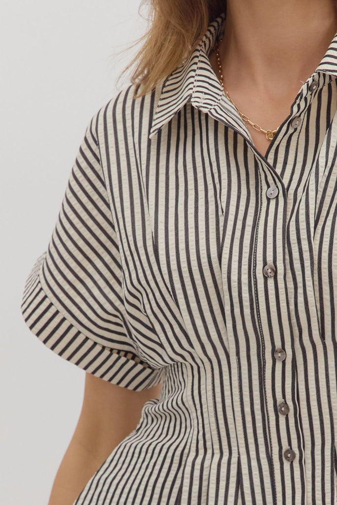 A woman wearing a vertical black and white striped short-sleeve blouse with a collar and button-down front, paired with tan shorts.
