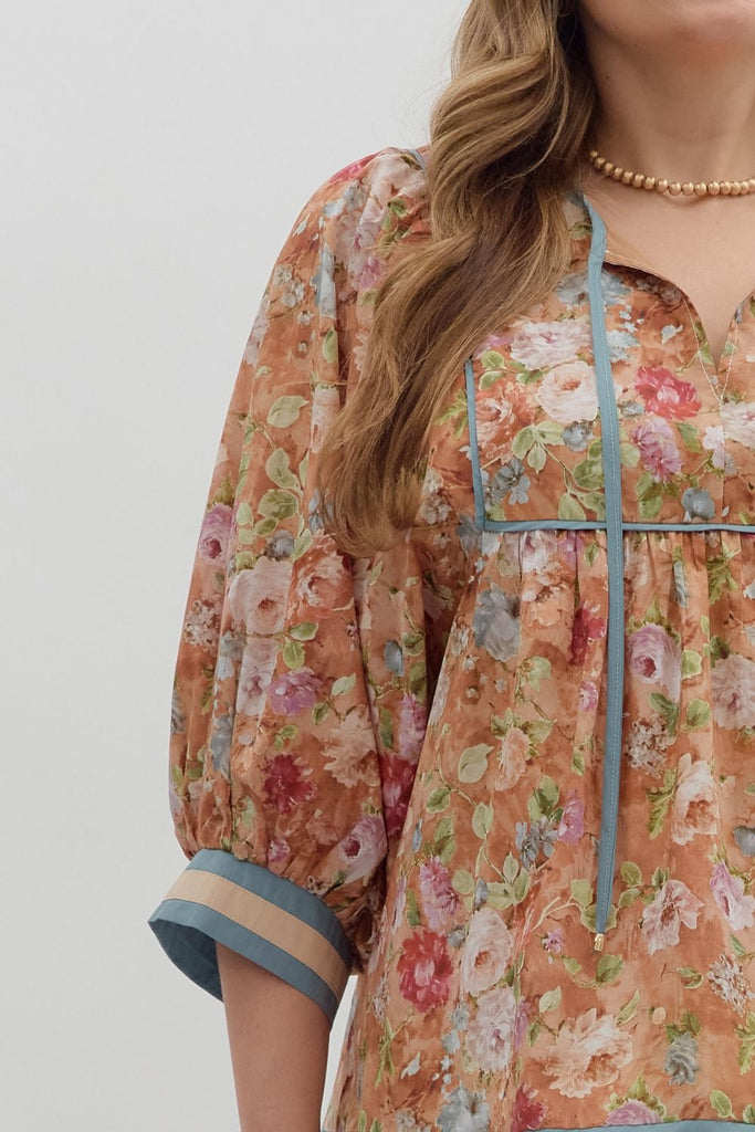 A woman wearing a loose-fitting floral blouse with puffed ¾ sleeves and an empire waist. The top has a vintage floral print in warm tones with blue and cream accent bands on the sleeves and hem.