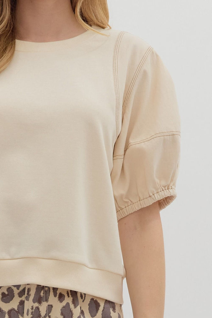 A woman wearing a beige crewneck top with short lantern sleeves and exposed seam details, paired with high-waisted leopard print shorts.
