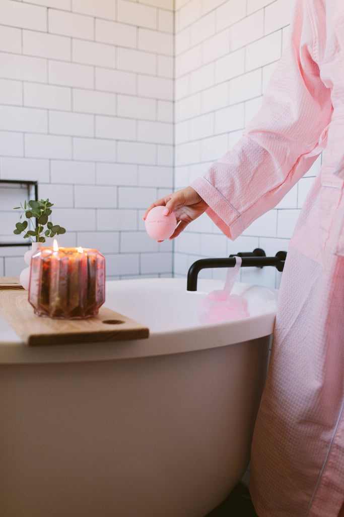 Person in a pink robe by a bathtub with a candle and plant, in a bathroom setting.