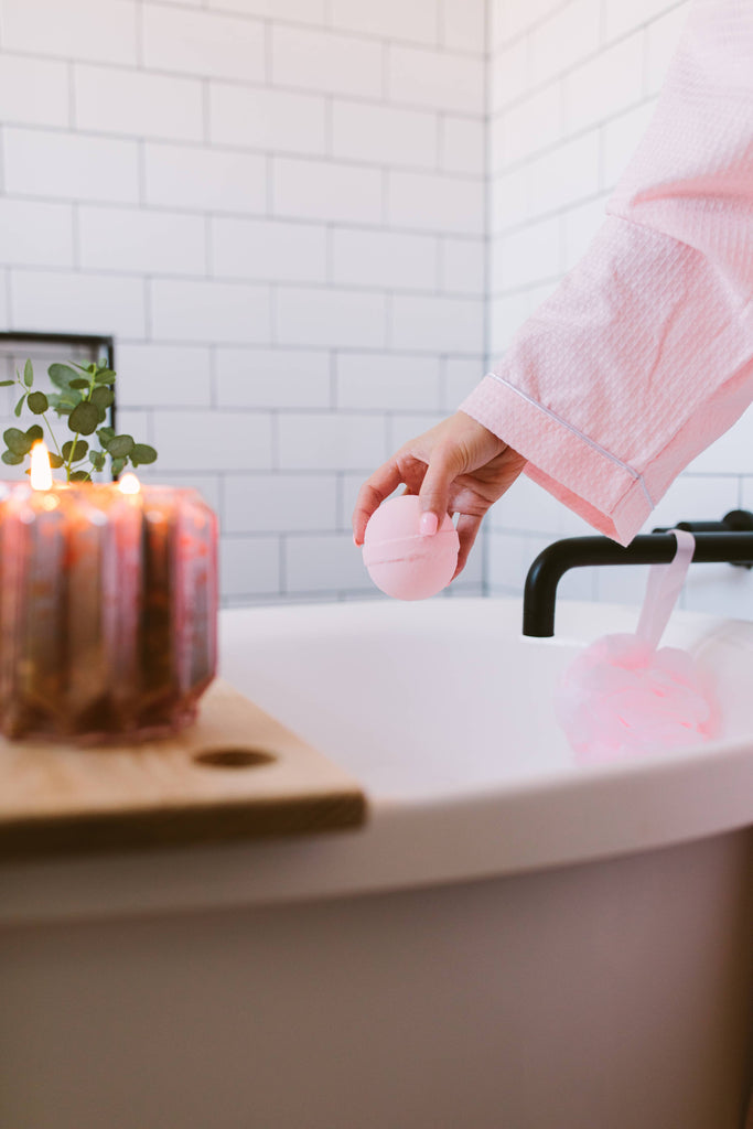 Person holding a pink bath bomb over a bathtub with candles and a plant in the background.