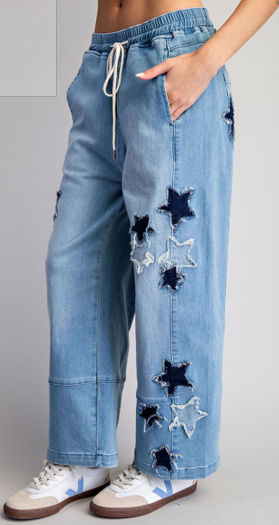 Close-up of light wash denim pants with raw-edged star appliqué patches, relaxed straight-leg jogger style.