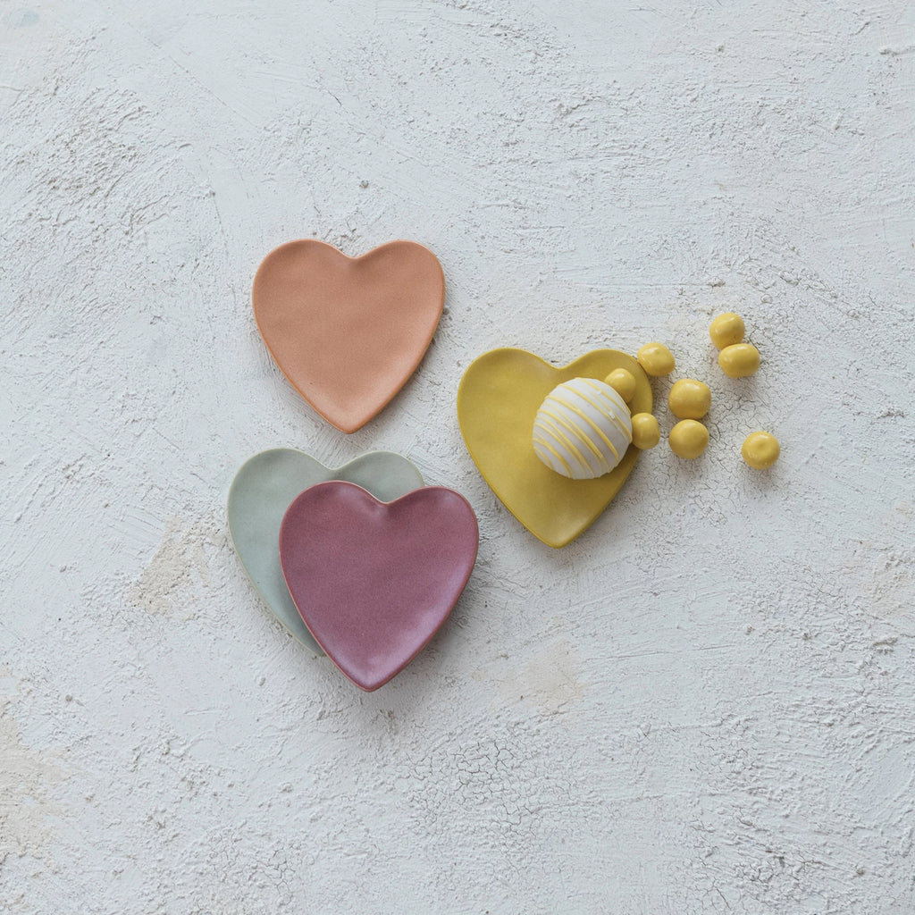 Four ceramic heart-shaped plates in different colors on a light gray surface with small yellow items.