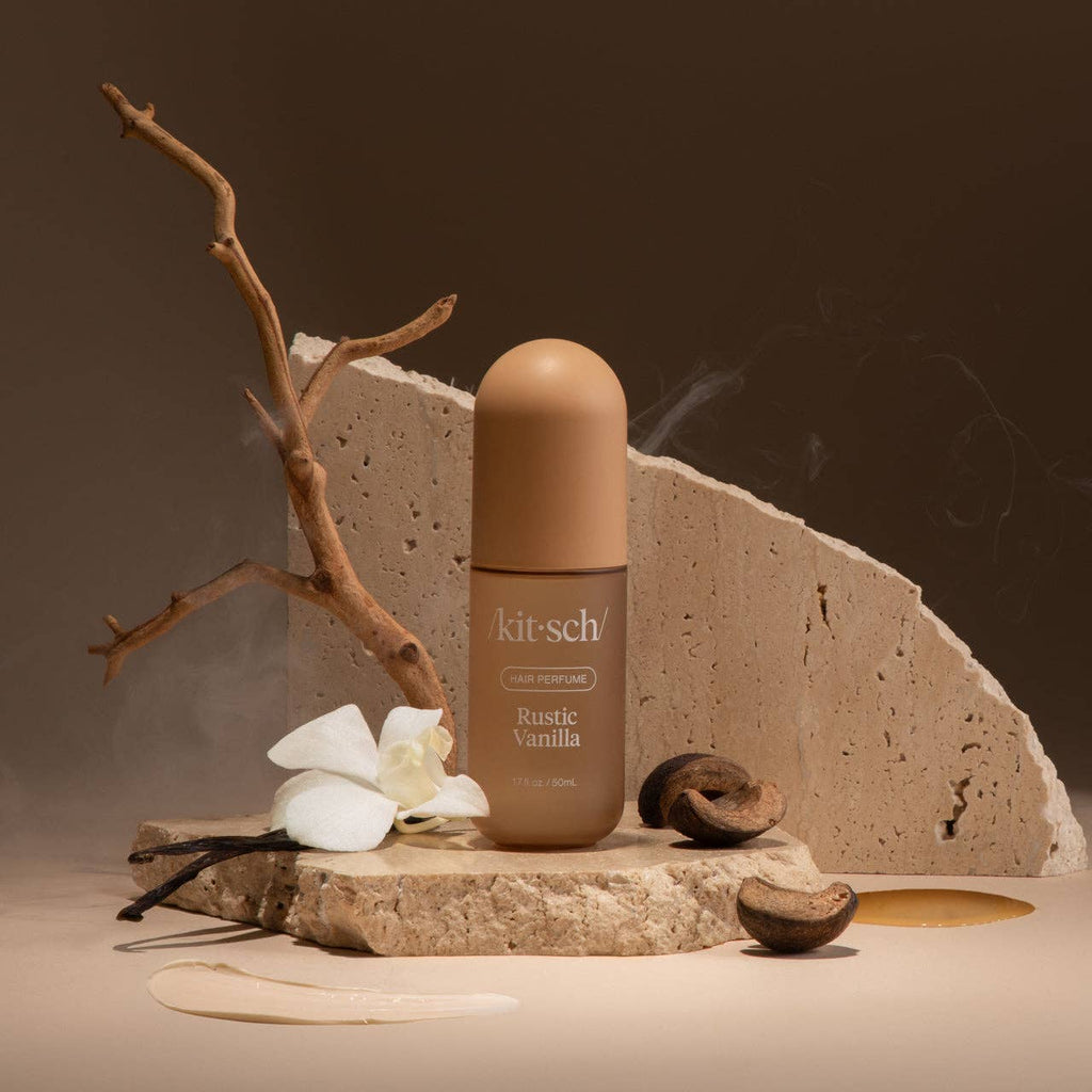 Kit-sch Rustic Vanilla perfume bottle on a stone surface with vanilla beans and flowers.