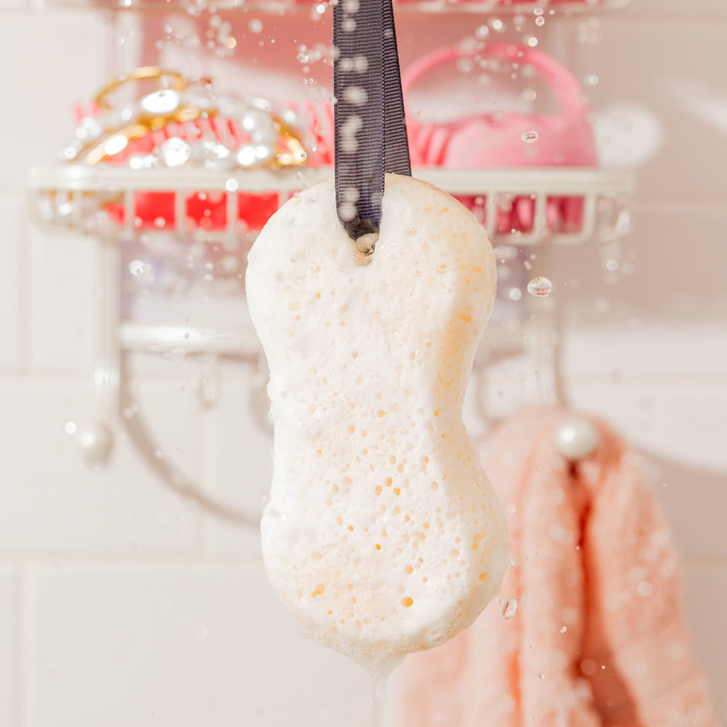 White bath bomb with gold specks hanging from a black ribbon against a blurred bathroom background.