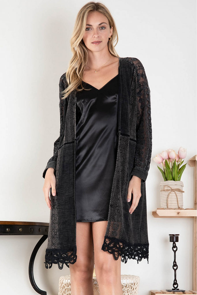 Woman wearing a black satin slip dress layered with a long black open-front cardigan featuring lace insets, front pockets, and scalloped lace hem, standing indoors with soft decor in the background.