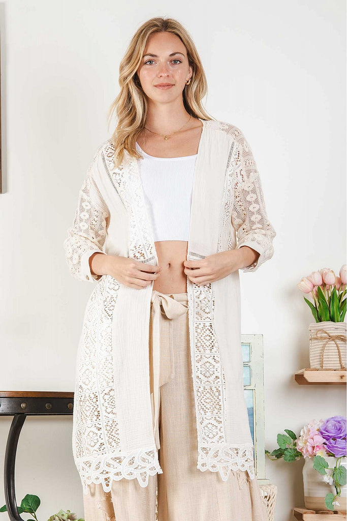 Woman wearing a natural-tone open cardigan with intricate white lace insets and scalloped hem, layered over a white ribbed crop top and beige wide-leg pants, standing indoors next to pastel floral décor.