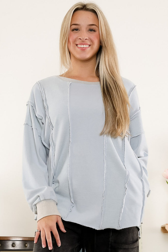Smiling woman wearing a light blue washed terry sweatshirt with exposed raw seams and ribbed cuffs, styled with black jeans in a casual indoor setting.