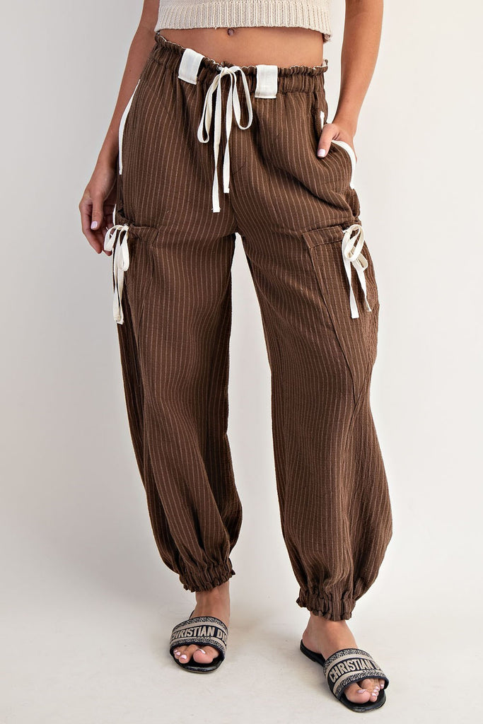 Model wearing relaxed-fit brown striped jogger pants with an elastic waistband and contrasting white drawstring ties. Pants feature utility-style flap pockets, front and back patch pockets, and gathered elastic hems, styled with open-toe logo slides.
