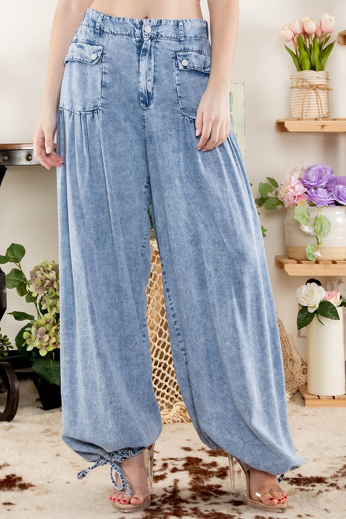 Close-up of a woman wearing high-rise, light-washed wide-leg denim pants with large snap-button patch pockets and adjustable drawstring cuffs, styled with clear high heels, standing indoors next to floral decor.