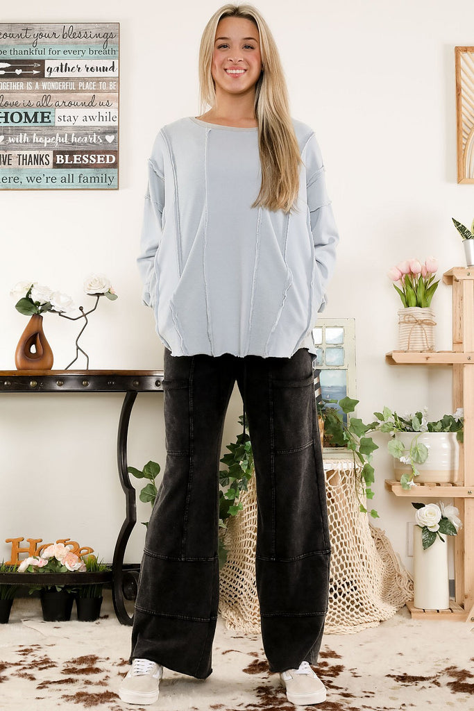 Smiling woman wearing a light blue washed terry sweatshirt with exposed raw seams and ribbed cuffs, styled with black jeans in a casual indoor setting.
