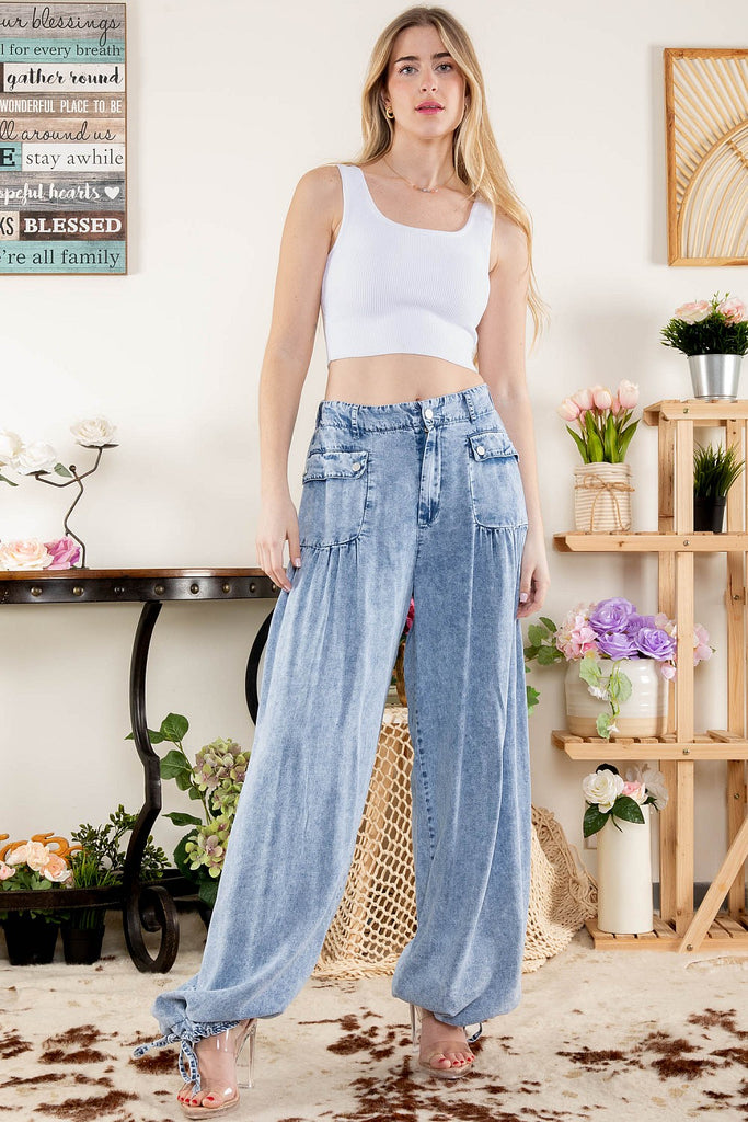Close-up of a woman wearing high-rise, light-washed wide-leg denim pants with large snap-button patch pockets and adjustable drawstring cuffs, styled with clear high heels, standing indoors next to floral decor.