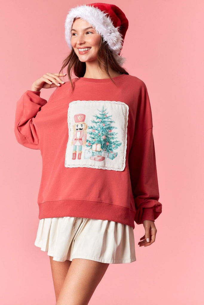 Dark coral Christmas sweatshirt featuring a nutcracker and Christmas tree corduroy patch with scalloped lace trim, styled with a pleated skirt and Santa hat.