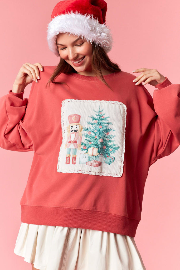 Dark coral Christmas sweatshirt featuring a nutcracker and Christmas tree corduroy patch with scalloped lace trim, styled with a pleated skirt and Santa hat.