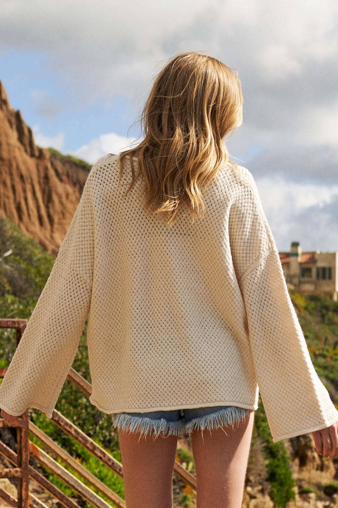 Person wearing a beige sweater and denim shorts standing on a cliff overlooking a scenic landscape.