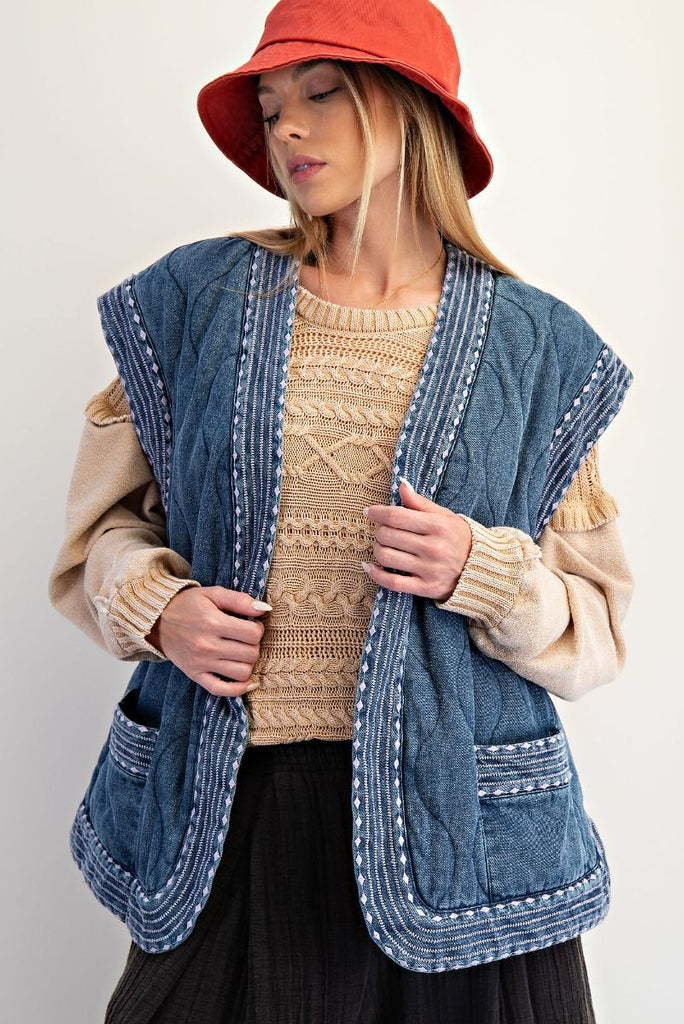 Woman wearing a quilted denim vest with embroidered trim and cap sleeves, styled over a beige knit sweater for a cozy boho look.