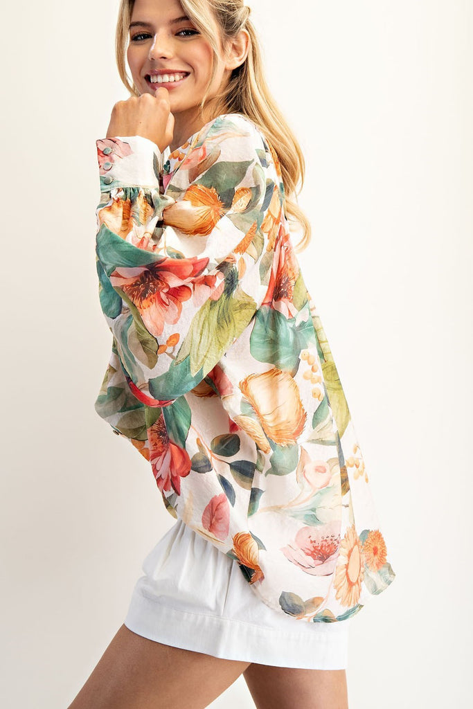 Model wearing a billowy, floral-printed blouse with balloon sleeves, a button front, and a high-low hem, styled with high-waisted white shorts.